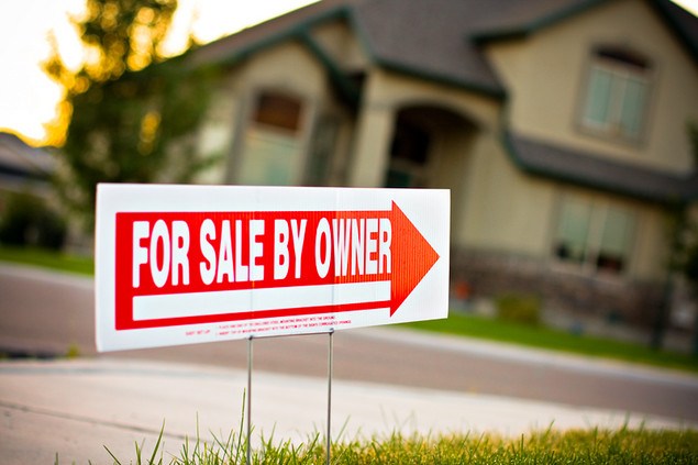 Brown and beige house in the background with red a white for sale by owner sign with a red arrow pointing towards the house.